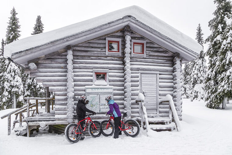 Kaksi pyöräilijää tutkimassa opastetauluja, jotka on kiinnitetty lumikuorrutetun hirsirakennuksen seinään. Tuvan ympärillä on kuusimetsää.