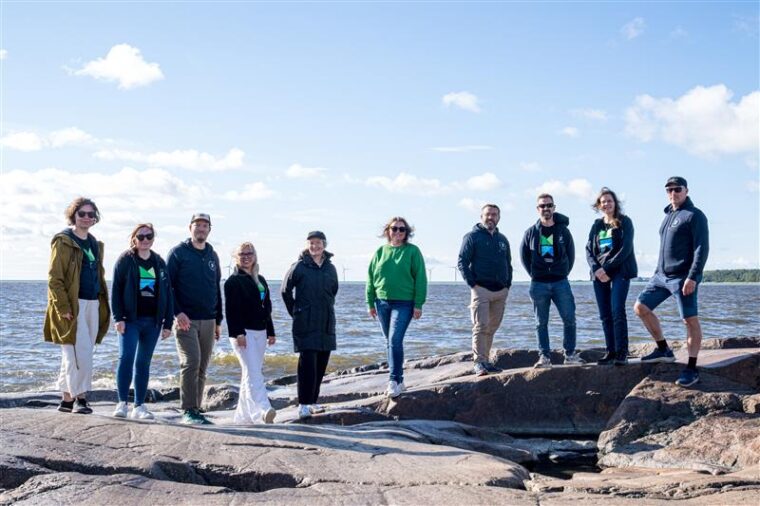 Kymmenen ihmist&auml; seisomassa aurinkoisella merenrantakalliolla meri taustalla.