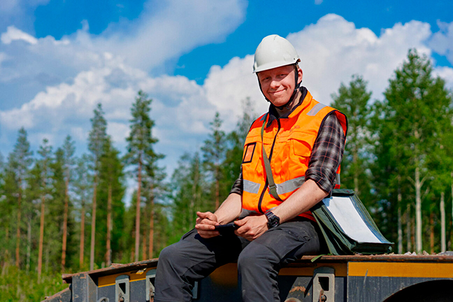 Henkilö kypärä päässään istuu jollakin korokkeella metsänreunassa ja hymyilee.