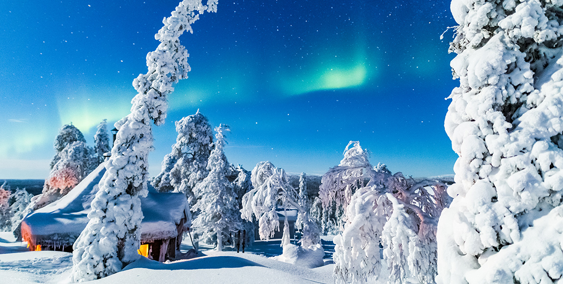 Lumista metsää ja mökki, jonka ikkunassa palaa valo. Taivaalla revontulia.
