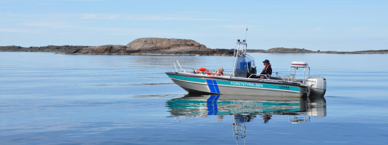 A Metsähallitus boat glides quietly through the archipelago with two people on board. The sea is calm, with rocky islands in the background.
