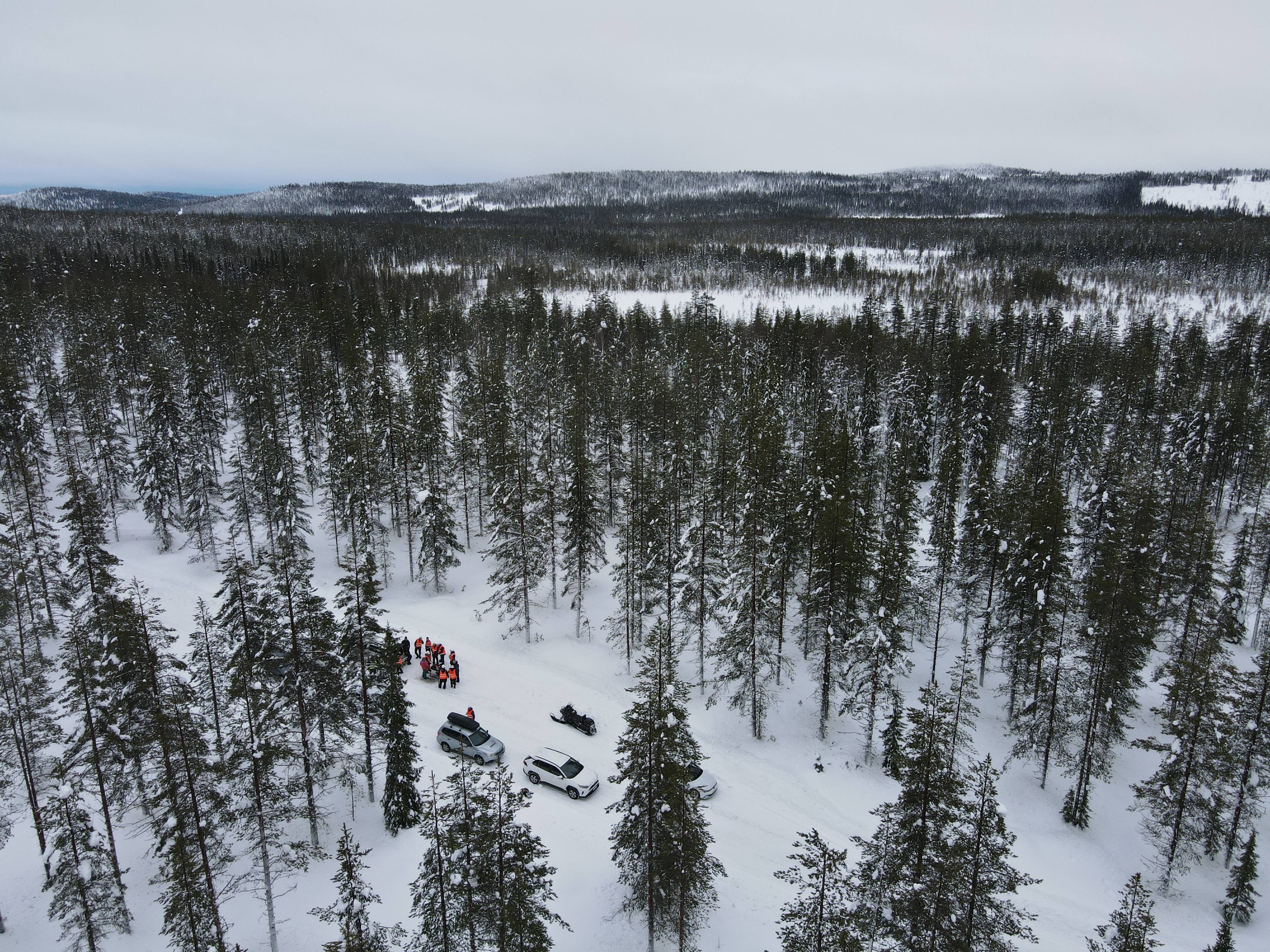 Ilmakuva lumisesta metsästä, maantasolla henkilöitä ja autoja.