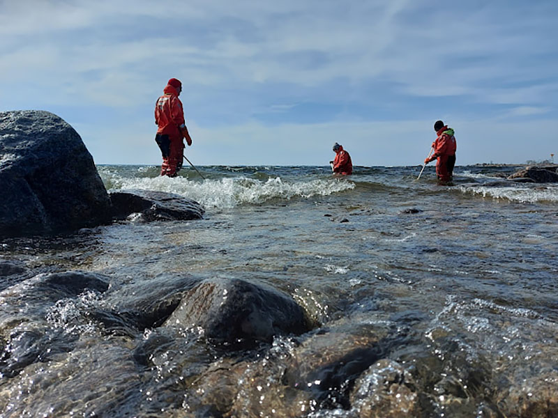 Kolme pelastautumispukuista henkilöä rantavedessä.