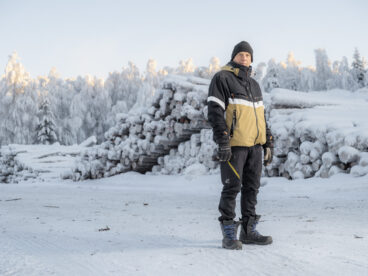 Henkilö seisoo lumisessa maastossa takanaan lumisia puupinoja.