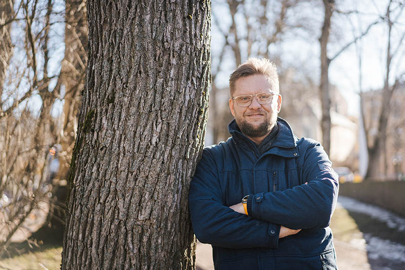 Vaaleahiuksinen silmälaseja käyttävä mies nojaa kookkaan puun runko vasten.