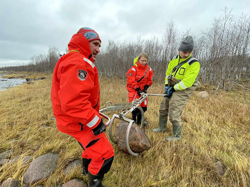 Kolme pelastautumispukuista henkilöä nostaa kiveä kivennostintyökalulla syksyisen saaren rannalla.