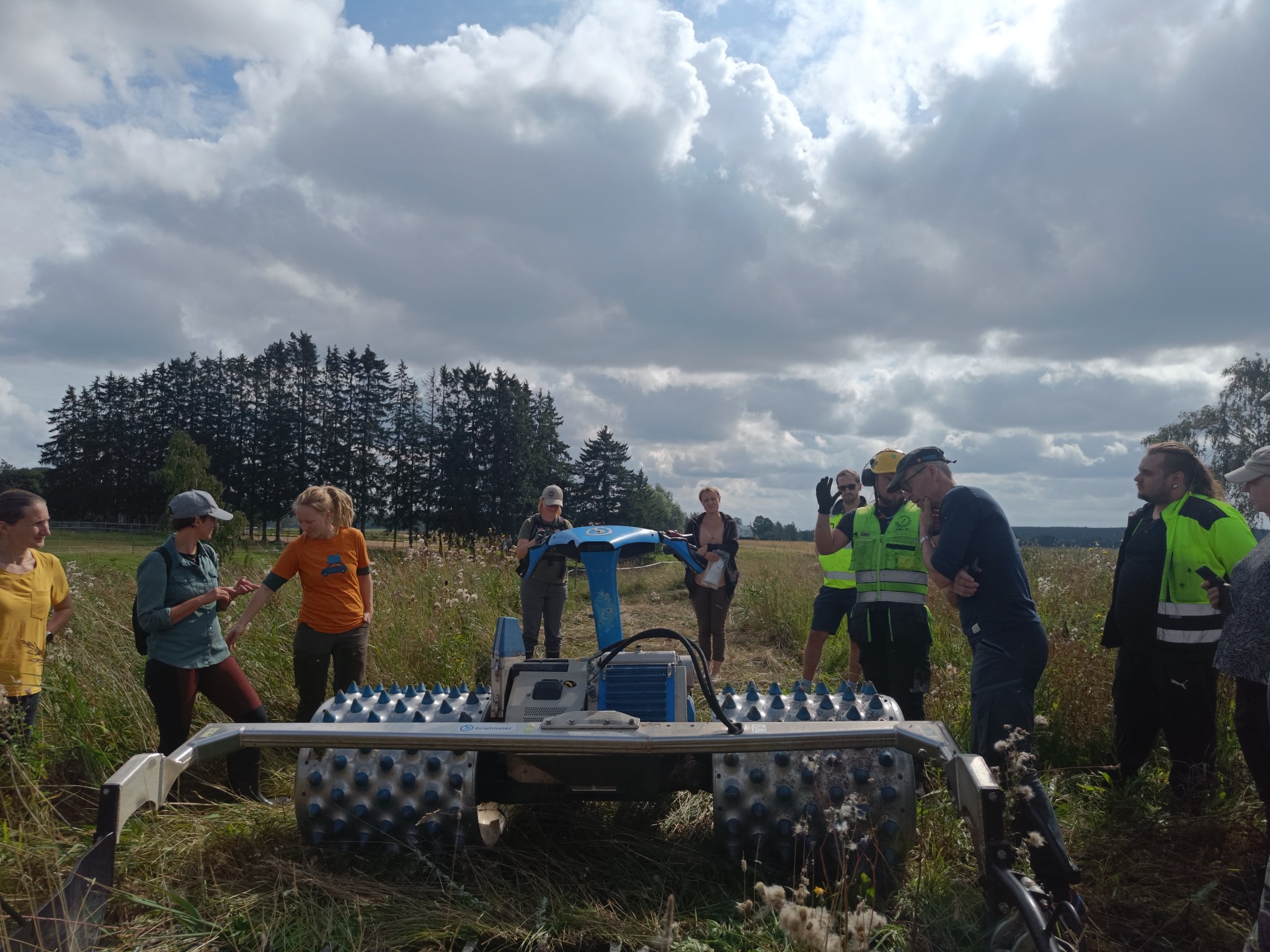 Ryhmä ihmisiä seisoo niittokoneen ympärillä niityllä.