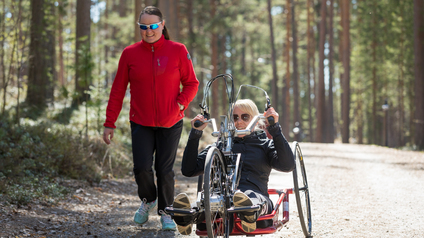 Kaksi henkil&ouml;&auml; kulkevat kes&auml;ll&auml; esteett&ouml;m&auml;ll&auml; reitill&auml;. Toinen kulkee k&auml;vellen, toinen kulkee urheilupy&ouml;r&auml;tuolilla.