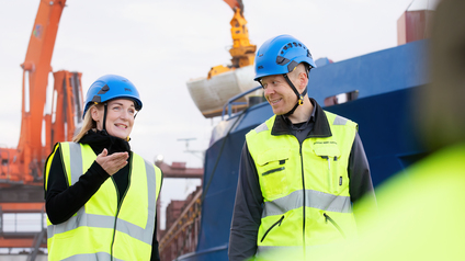 Kaksi henkil&ouml;&auml; keltaisissa huomioliiveiss&auml; ja siniset kyp&auml;r&auml;t p&auml;&auml;ss&auml; k&auml;velee satamassa.