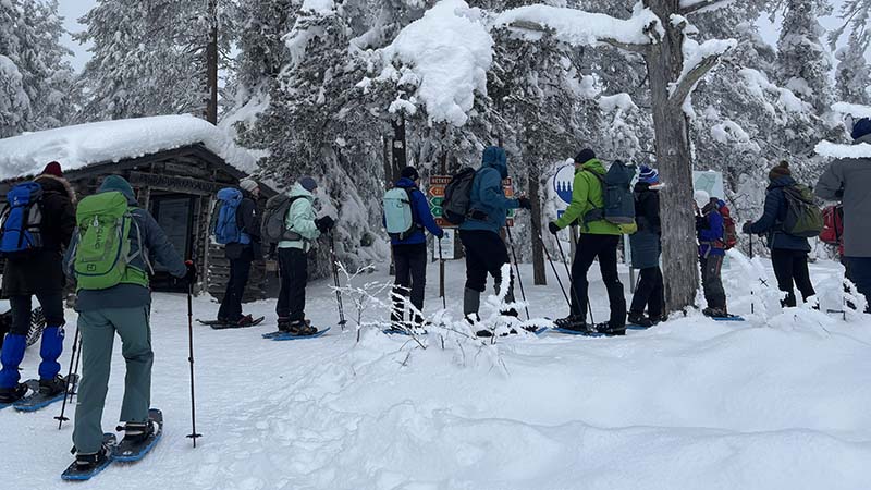 Lumikenkäilijöitä viittojen, auton ja pienen hirsisen rakennuksen luona.