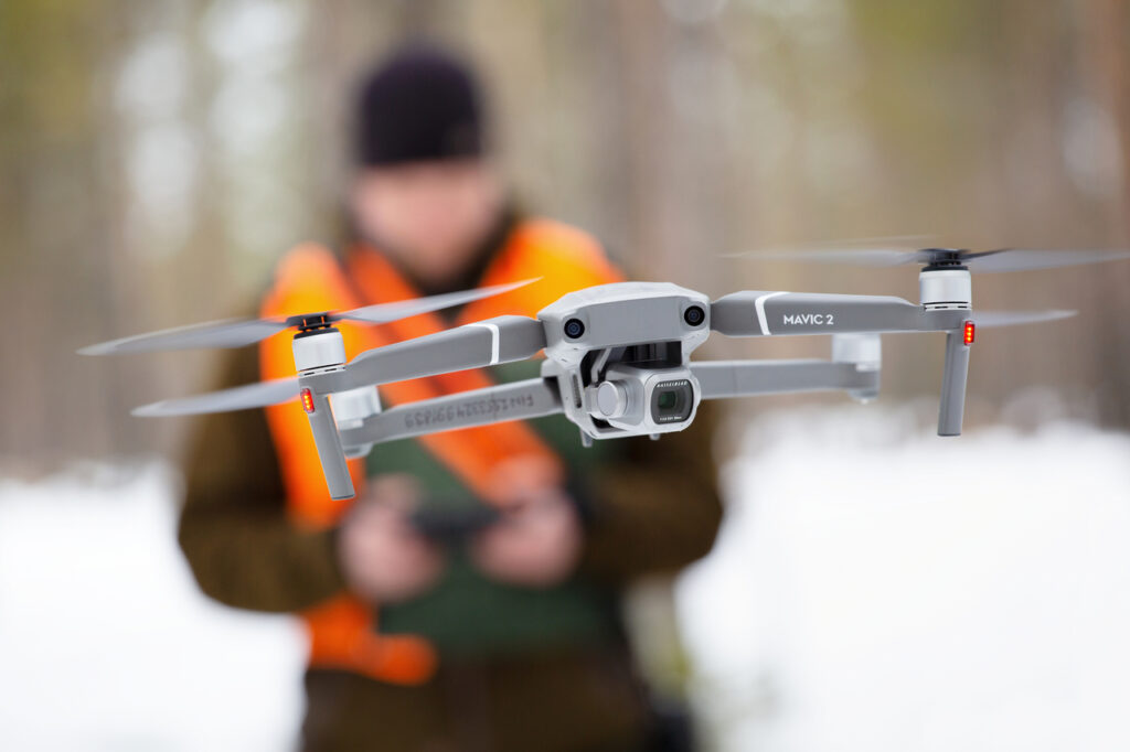 Lähikuva ilmassa lentävästä droonista. Oranssiliivinen suunnittelija taustalla epätarkkana.