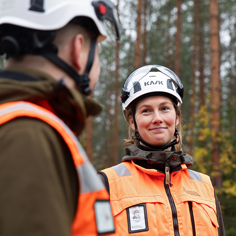 Kaksi henkilöä metsässä kypärät päässä. Toinen on kameraan päin selin ja toinen katsoo selin olevaa.