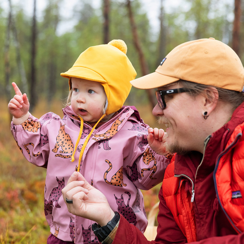 Pieni lapsi ja mies syksyisessä metsässä. Lapsella on sadetakki ja mies on kyykyssä lapsen vieressä.