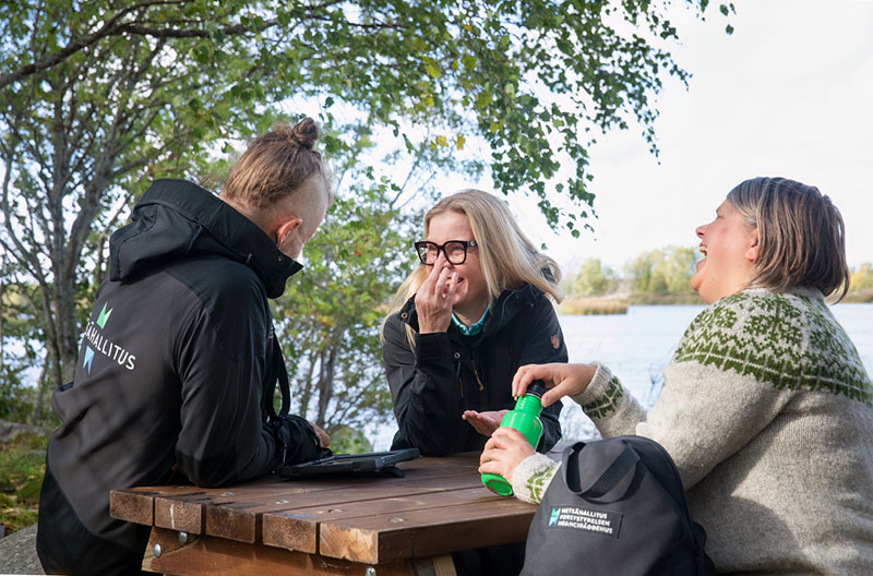Kolme henkilöä istuu ulkona puisen pöydän ääressä retkivaatteissa ja juttelee nauraen.