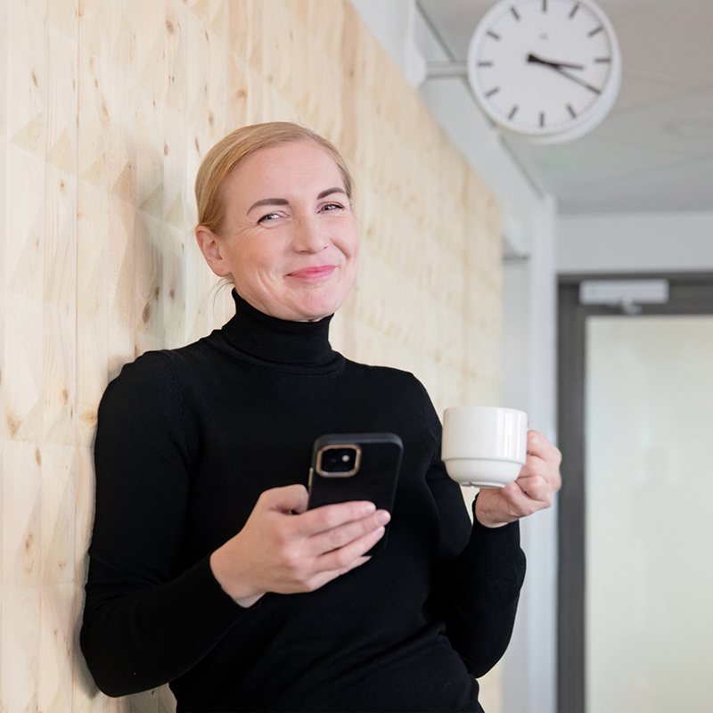 Hymyilevä nainen nojaa toimiston seinään ja hänellä on toisessa kädessä kahvikuppi ja toisessa kännykkä.