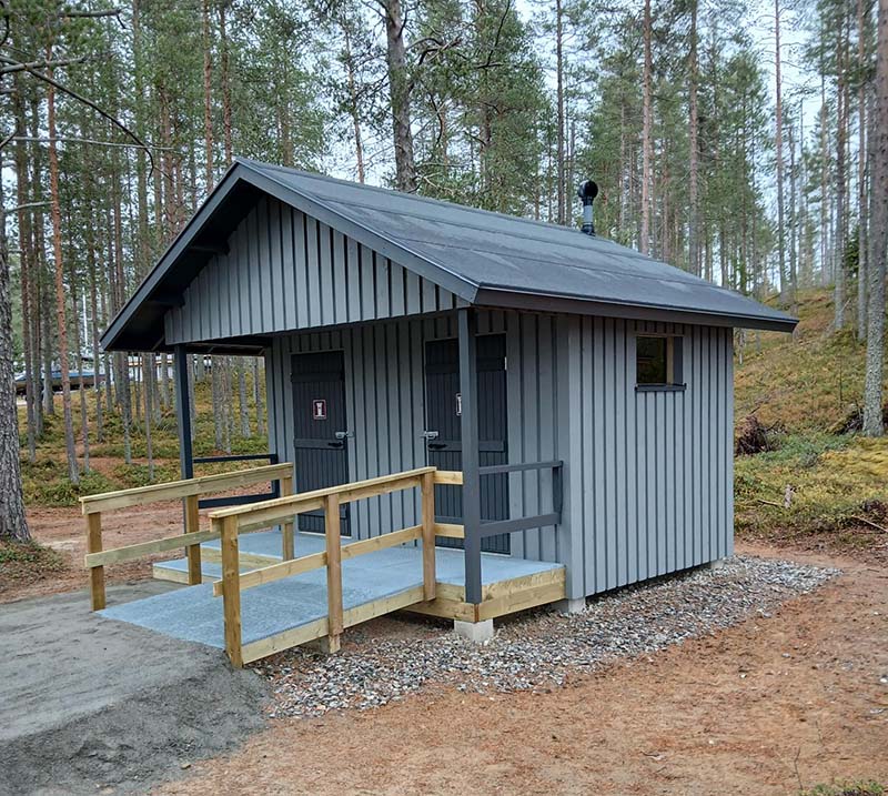 Puinen kuivakäymälärakennus, jossa on yksi esteetön ja yksi ei-esteetön käymäläosasto. Ovien edessä on tasanne ja luiska teräsritilästä sekä puiset kaiteet.