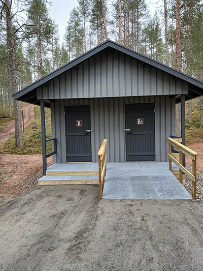 Puinen kuivakäymälärakennus, jossa on yksi esteetön ja yksi ei-esteetön käymäläosasto. Ovien edessä on tasanne ja luiska teräsritilästä sekä puiset kaiteet.