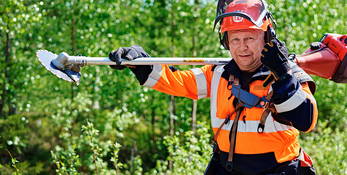 A man dressed in an orange safety suit with a chainsaw on his shoulder. A lush forest behind.