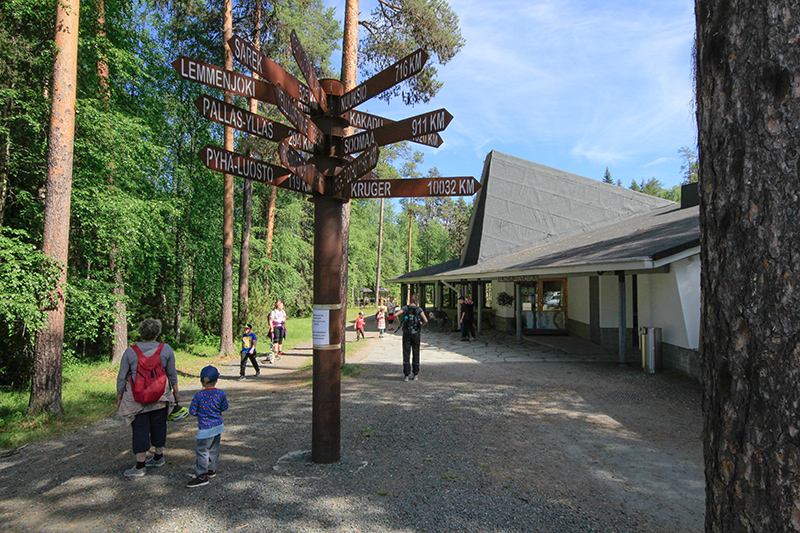 Rakennuksen pihalla on ihmisiä kesällä. Keskellä pihaa on iso viittaopaste.
