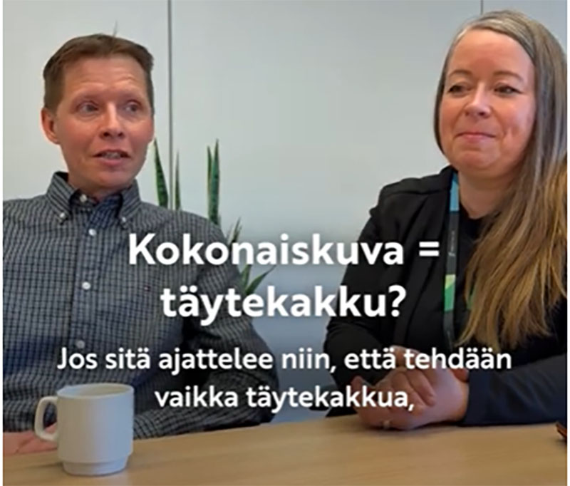 Kaksi henkilöä istuu pöydän ääressä ja toisen edessä on muki. Henkilöiden päällä on teksti: "Kokonaiskuva täytekakku? Jos sitä ajattelee niin, että tehdään vaikka täytekakkua,"