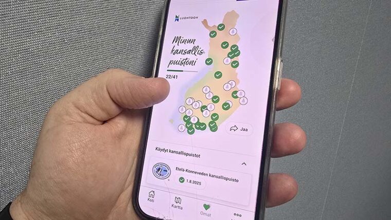 Käsi pitelee kännykkää, jonka näytöllä näkyy Suomen kartta sekä tekstit Luontoon, Minun kansallispuistoni 22/41 sekä Etelä-Konneveden kansallispuisto 1.8.2025.