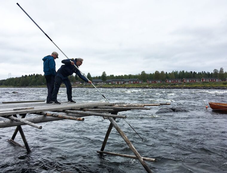 Kaksi miest&auml; seisoo laiturilla. Toinen pitelee pitk&auml;&auml; keppi&auml;, jonka toinen p&auml;&auml; on vedess&auml;.