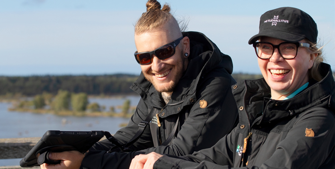 Kaksi hymyilevää henkilöä seisovat lintutornissa ja nojaavat kaiteeseen. Toisella on kädessään kannettava tietokone.