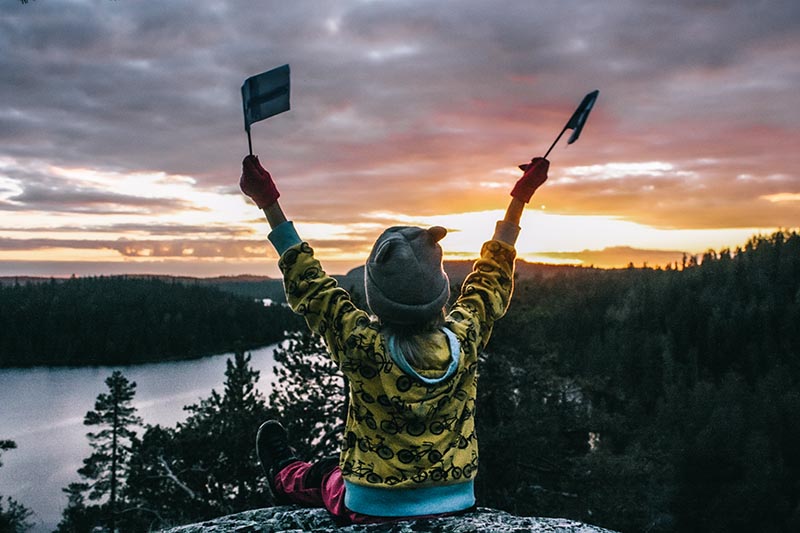 Kuvassa etualalla kalliolla istuva lapsi pienet Suomen liput käsissään. Lapsi katsoo kamerasta pois päin auringonlaskua. Taustalla järvimaisema ja metsää.
