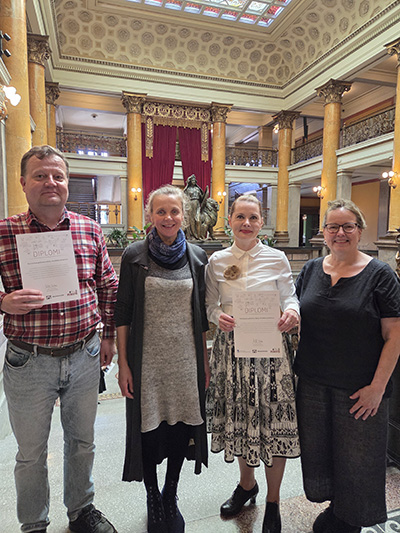 Neljä henkilöä seisoo juhlavassa tilassa ja katsoo kohti kameraa. Kahdella on käsissään diplomi.