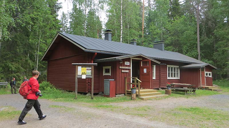 Iso hirsinen kämppärakennus kahdella oviaukolla. Kesäisessä maisemassa etualalla retkeilijä.