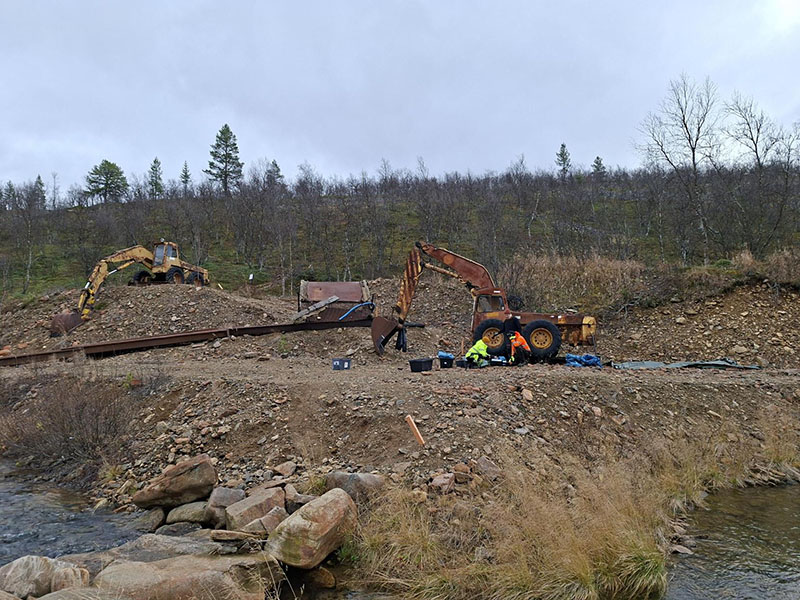 Kaksi kaivinkonetta. Toisen kaivinkoneen vieressä on kyykyssä kaksi henkilöä. Heidän ympärillään on lainapeite, muovilaatikoita ja säkkejä.