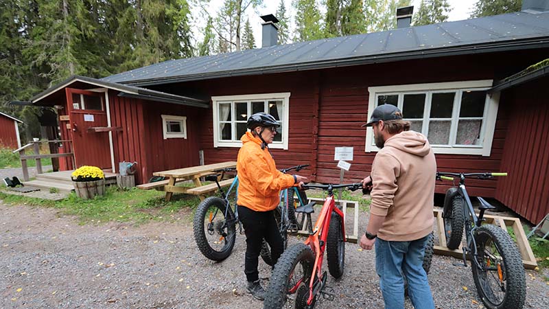 Kaksi henkilö hirsirakennuksen edustalla olevien pyörien luona. Toisella on pyöräilykypärä päässään.
